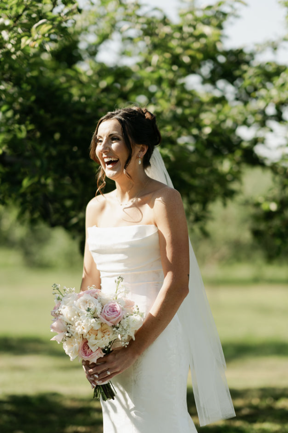 A smiling bride holding a bouquet of white and pink roses outdoors with trees in the background.