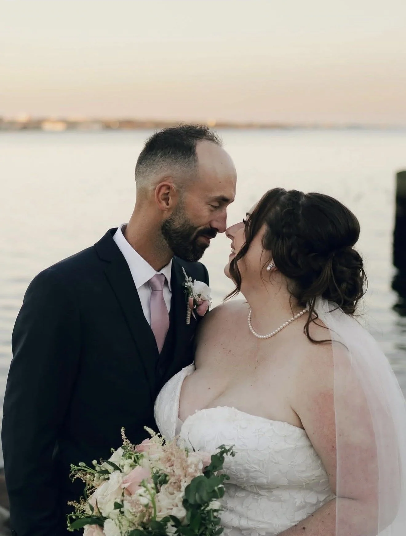 A newlywed couple shares an intimate moment by a body of water during sunset, with the groom in a dark suit and pink tie, and the bride in a white strapless wedding gown holding a bouquet of pink and white flowers.