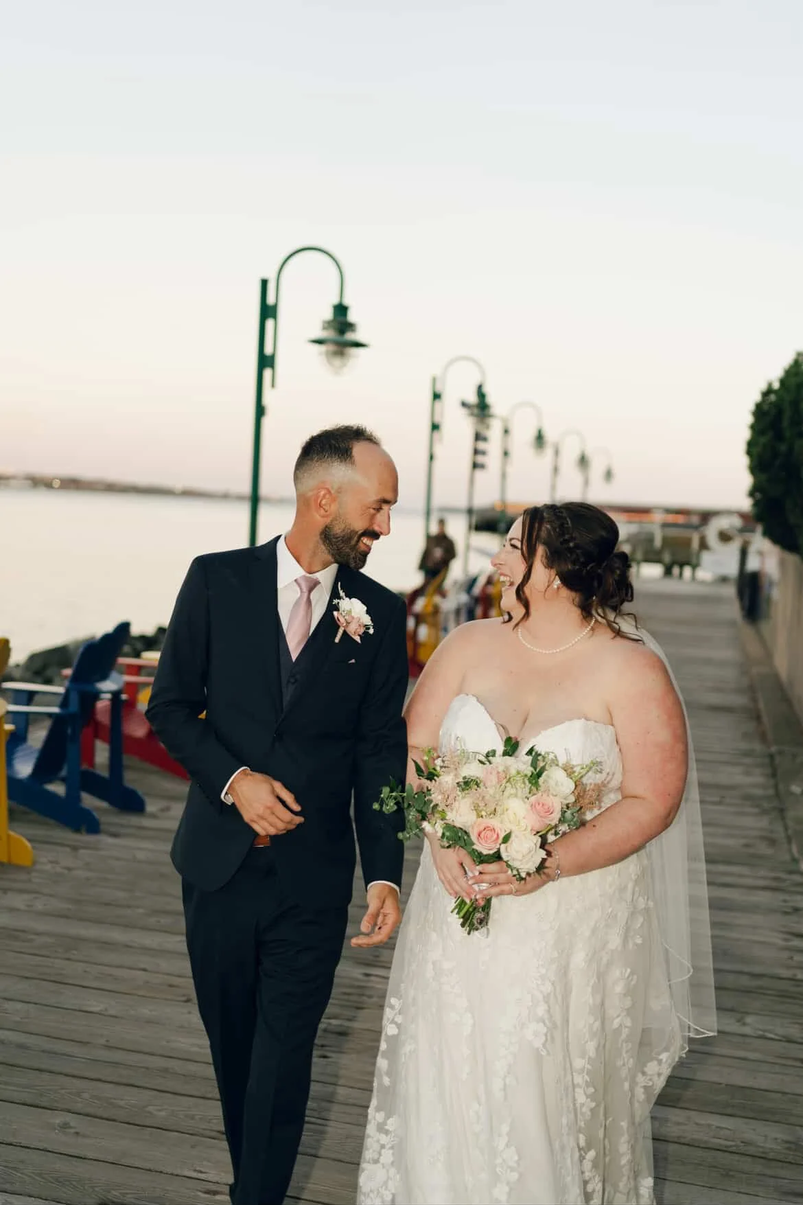 A newlywed couple walking by the water on a wooden pier at sunset, smiling at each other. The groom wears a black suit with a light pink tie and a boutonniere, and the bride wears a strapless white wedding gown with lace details, holding a bouquet of pink and white roses. String lights line the pier in the background.