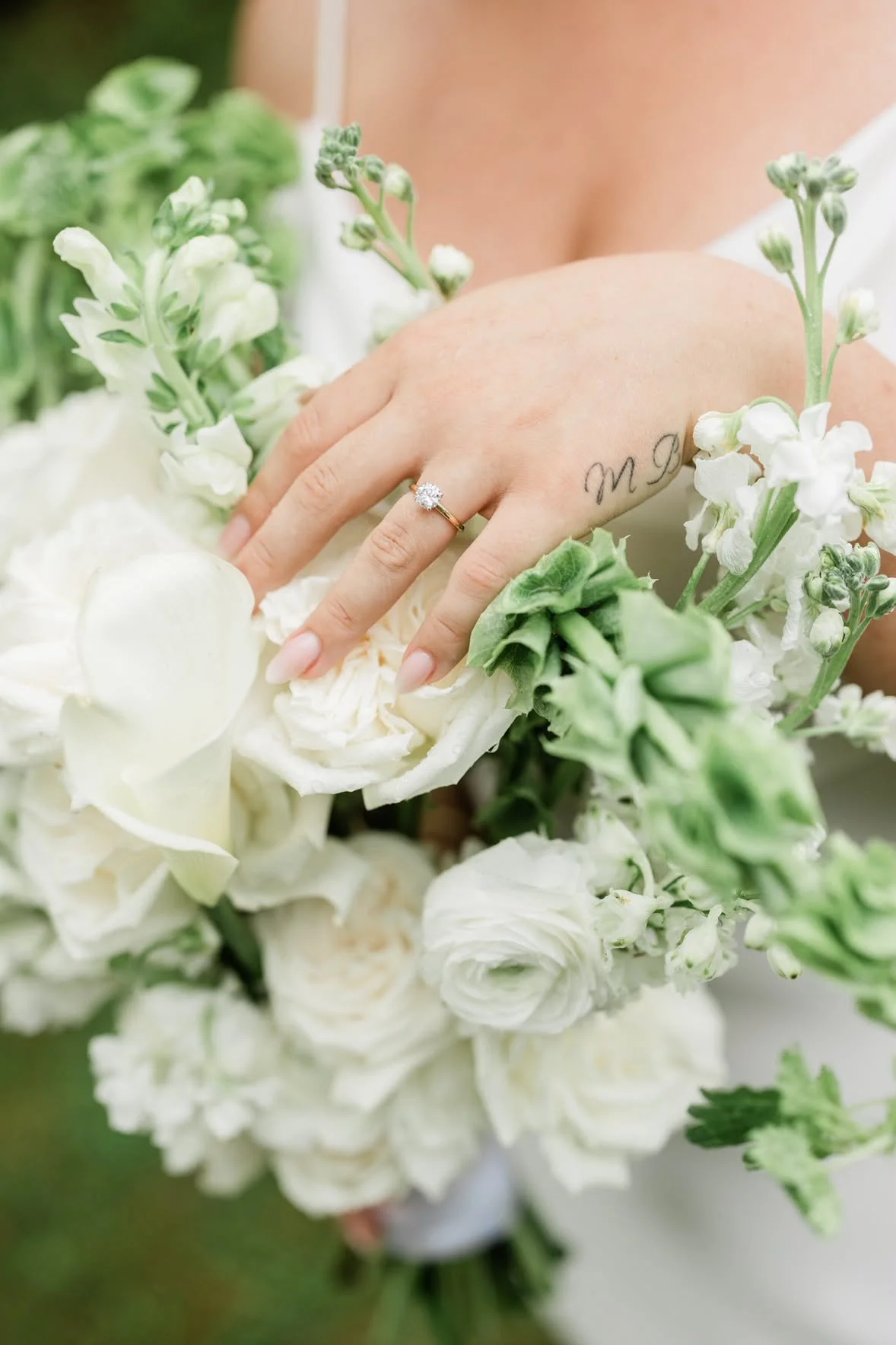 Close-up of a woman's hand with a wedding ring, surrounded by white and green flowers. The woman has a tattoo on her ring finger.