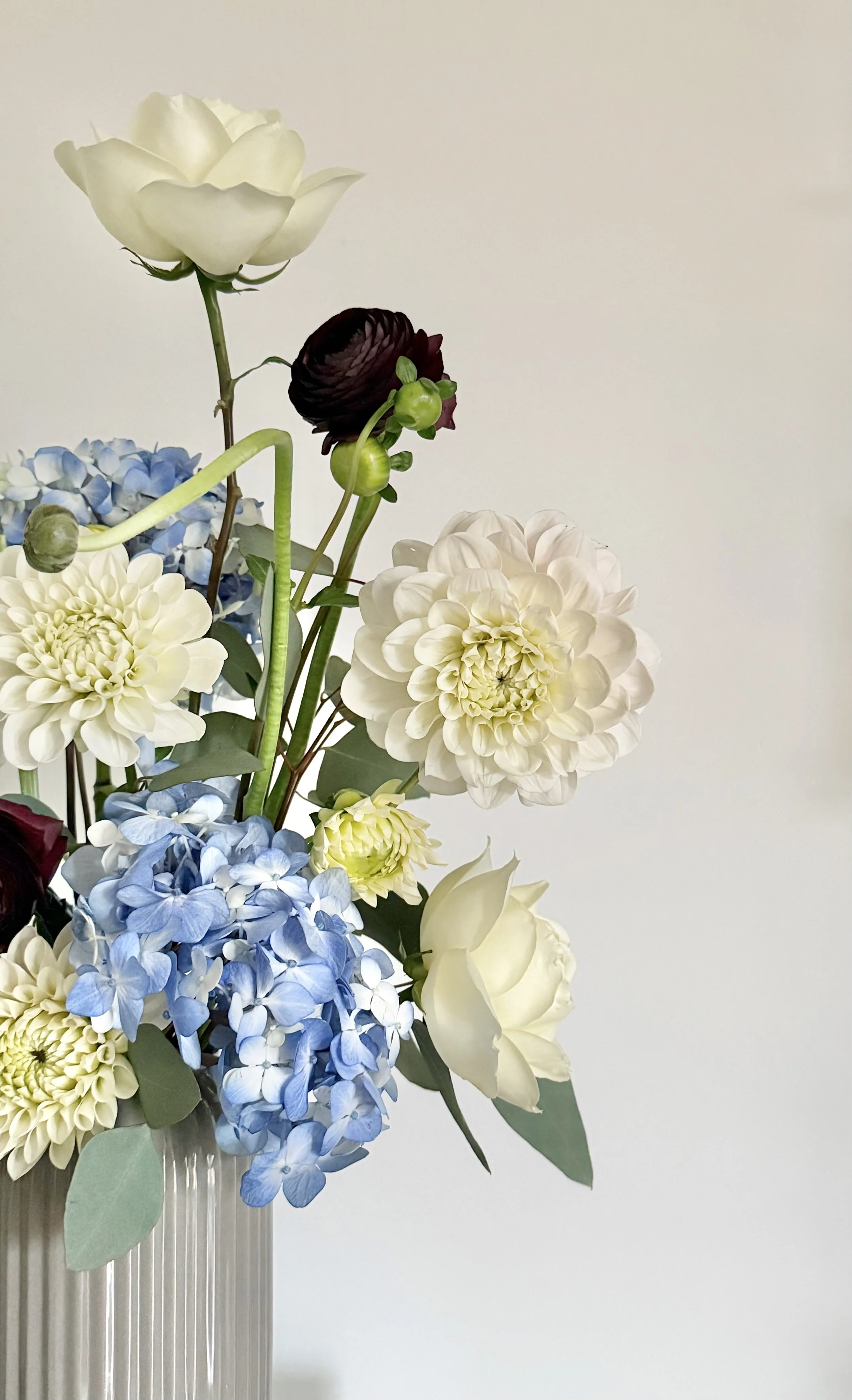 A bouquet of white, yellow, blue, and dark red flowers in a clear glass vase against a plain off-white background.