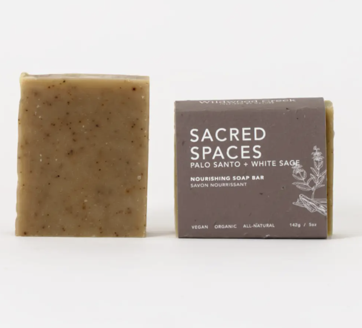 A bar of soap labeled "Sacred Spaces," featuring a minimalist design and earthy colors, resting on a wooden surface.