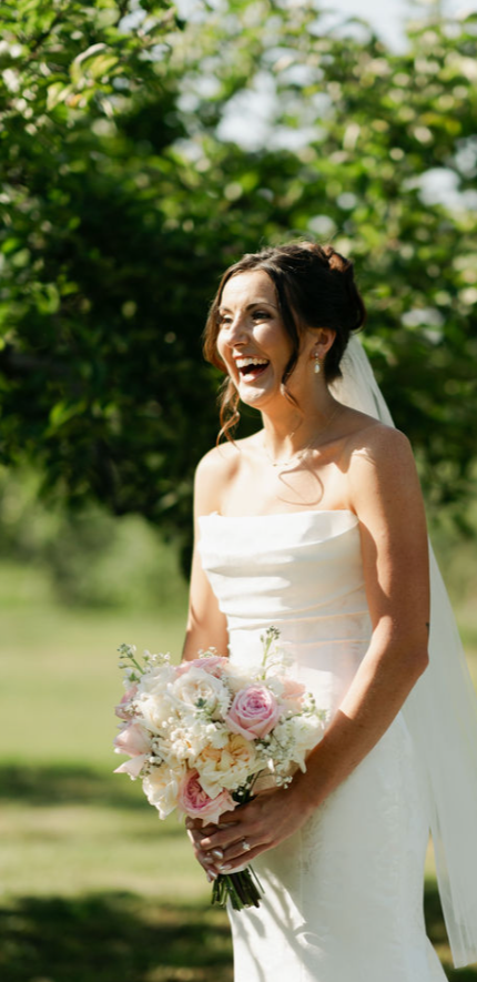 A bride with dark hair in an updo, wearing a strapless white wedding dress and holding a bouquet of white and pink flowers, standing outdoors with green trees in the background.
