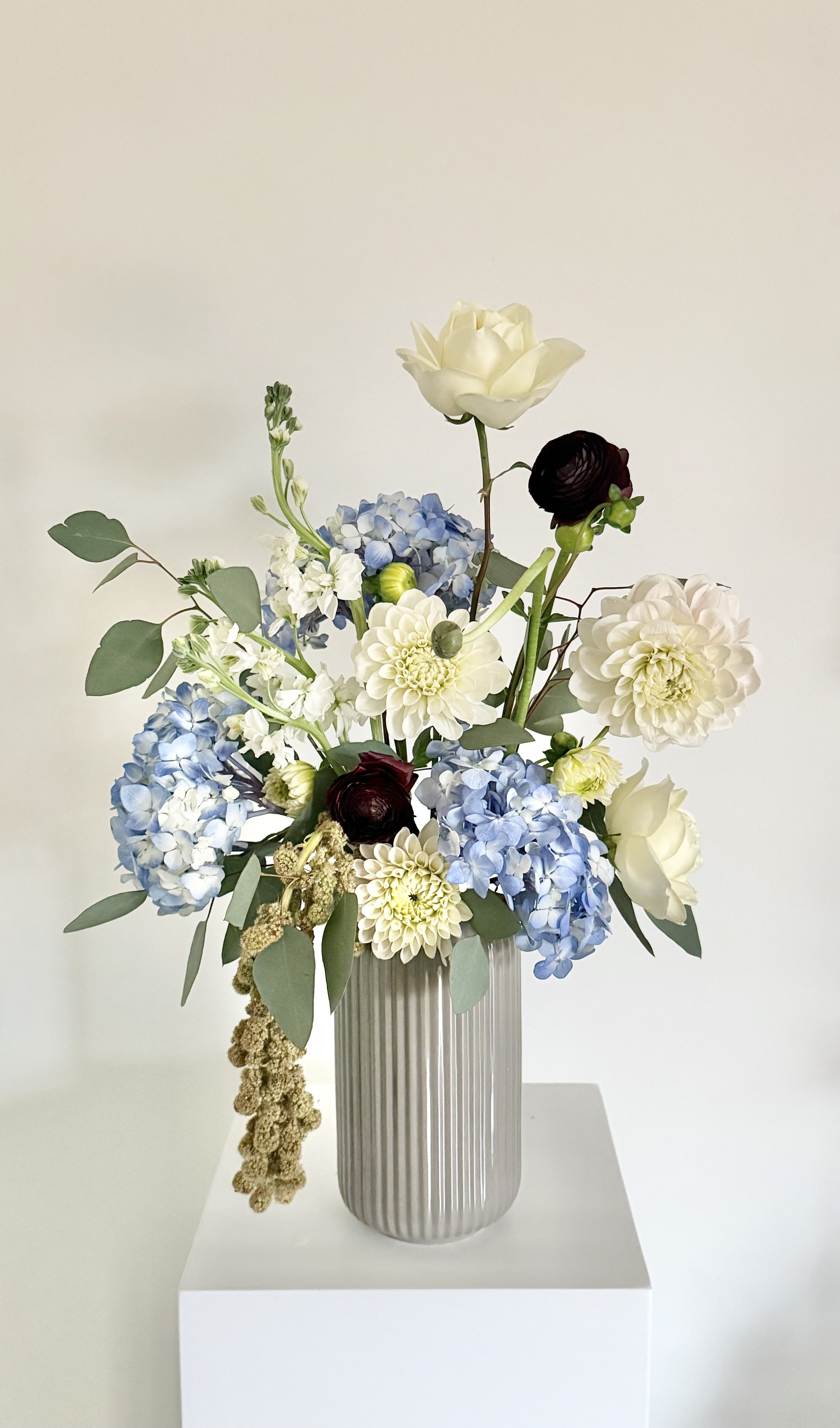 A floral arrangement with white, blue, and dark red flowers in a gray ribbed vase on a white pedestal against a plain white wall.