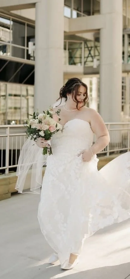 Bride holding a bouquet of white and pink flowers, standing outside in a modern urban setting.