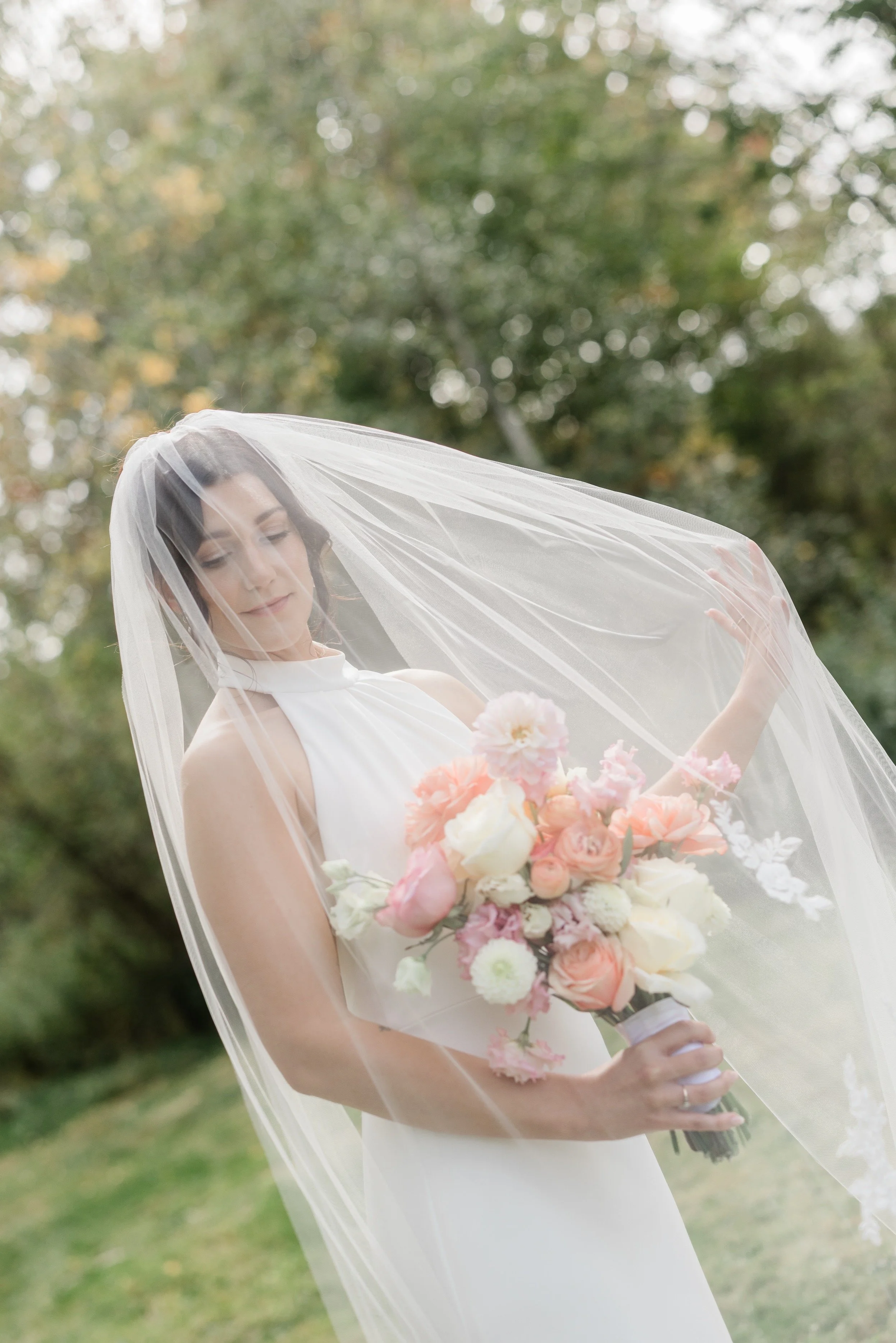 A bride outdoors holding a bouquet of pink and white flowers, wearing a white dress and veil, with her eyes closed and her head slightly tilted.