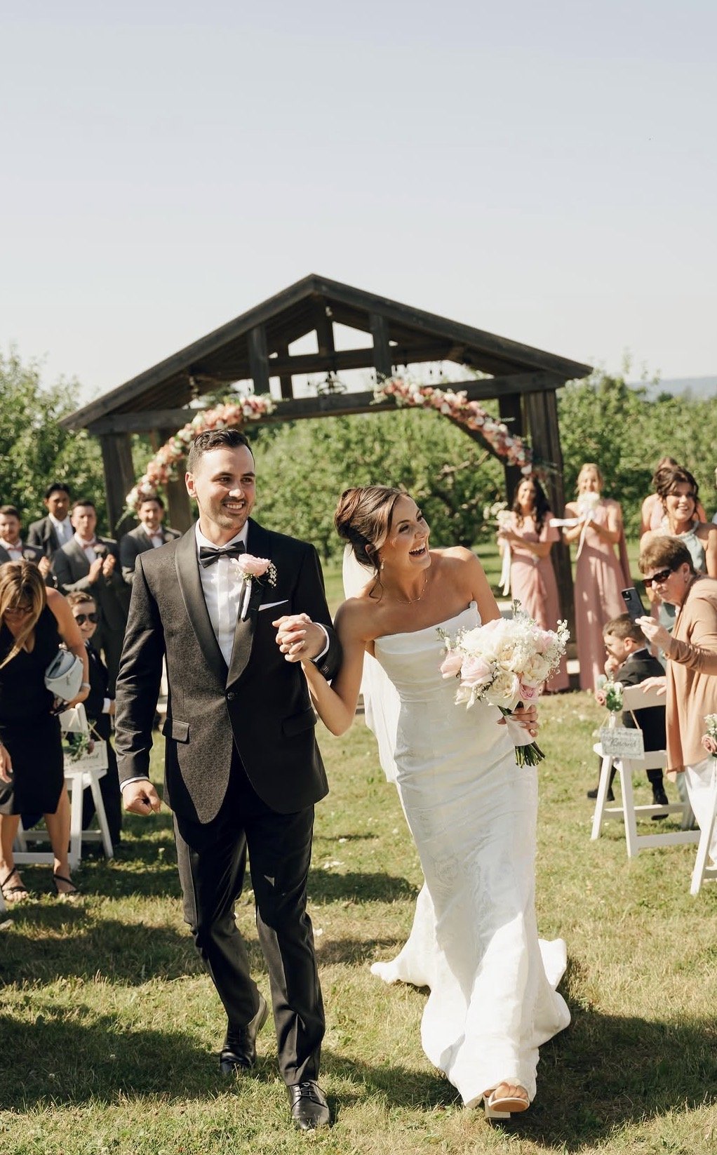 A newlywed couple walking down the aisle at an outdoor wedding ceremony, smiling and holding hands, with guests and bridesmaids in the background.