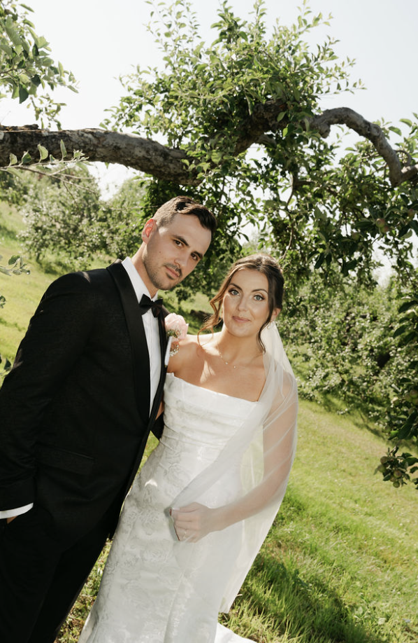 A newlywed couple in formal wedding attire stands outdoors under a tree, with green grass and foliage around them, during daytime.