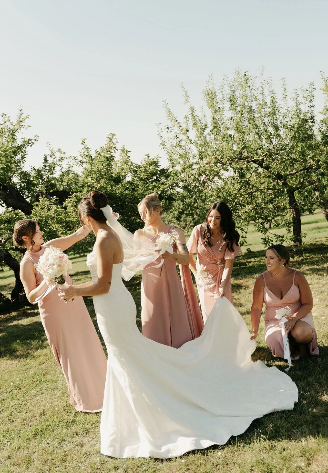 A bride in a white wedding dress is holding a bouquet and is surrounded by five women wearing blush pink dresses, standing outdoors in a grassy area with trees in the background.