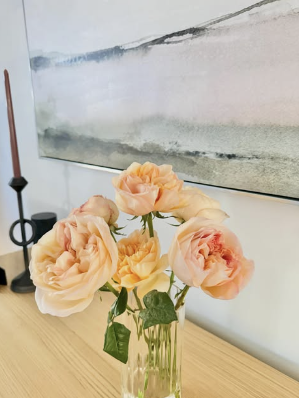 A clear glass vase with five peach-colored roses on a wooden surface. In the background, part of a painting with soft pastel colors and a candlestick with a pinkish candle is visible.