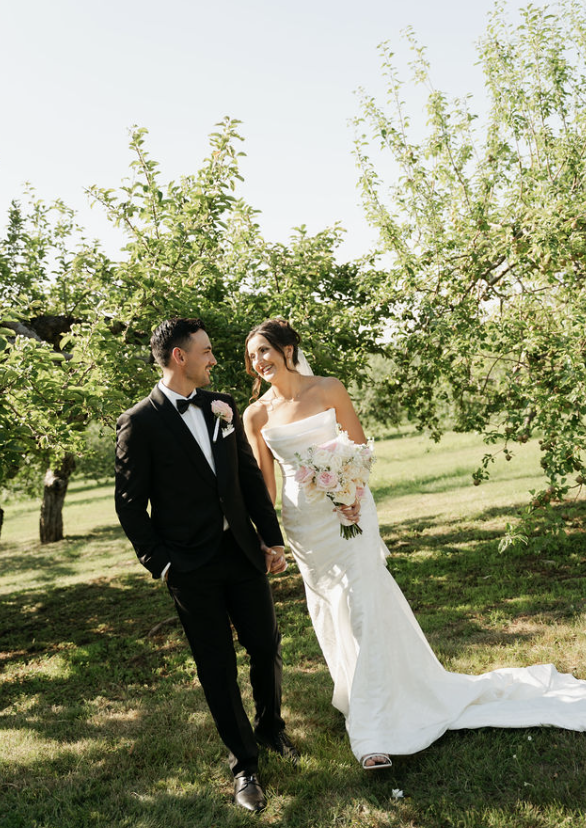 Bride and groom holding hands and smiling in a lush outdoor garden, with green trees and sunlight.