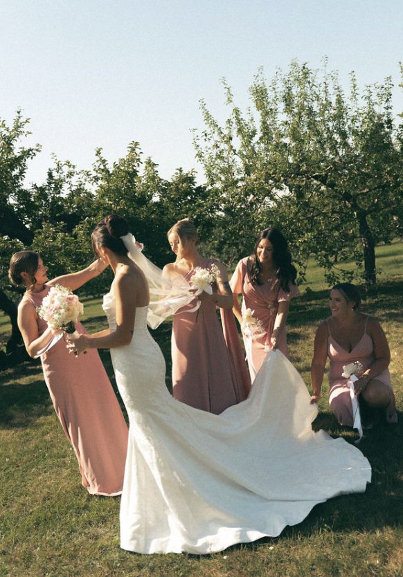 A bride in a white wedding dress and veil standing outdoors in a grassy area surrounded by four women in pink dresses, holding bouquets and assisting her. Trees are in the background, and the scene is sunny.