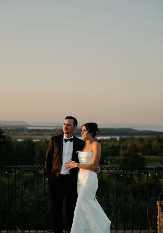 Bride and groom standing together outdoors at sunset, with a scenic landscape in the background, wedding attire, and a black metal fence.