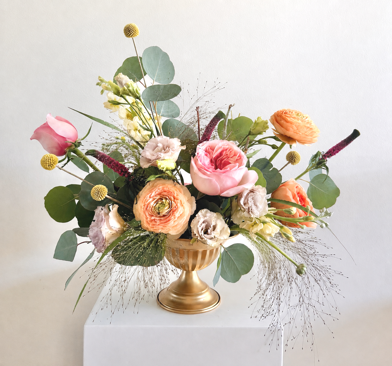 A floral arrangement with pink, peach, and white flowers in a gold vase on a white pedestal.
