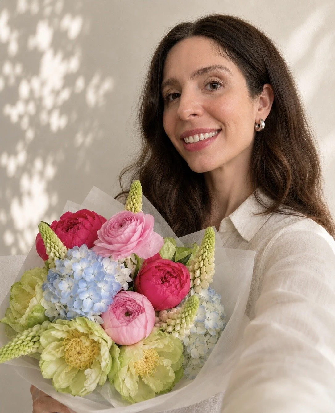 A woman with long brown hair, wearing a white shirt and silver hoop earrings, smiling and holding a colorful bouquet of flowers.