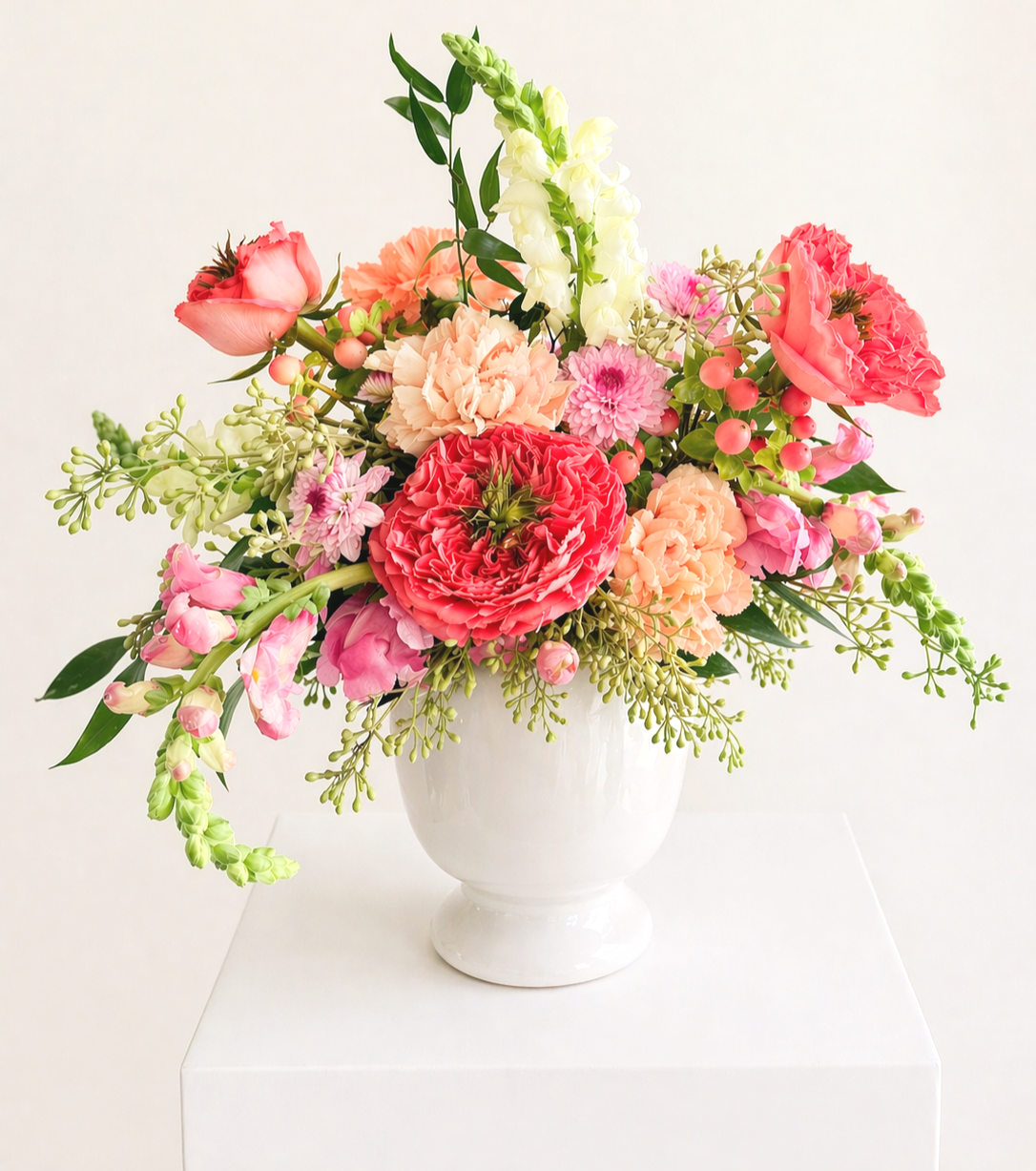 A bright and colorful mixed floral bouquet in a white vase on a white surface, featuring pink, peach, and red flowers with green foliage.