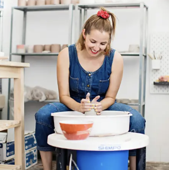 A woman sits at a potter's wheel, shaping clay with her hands in a creative workspace.