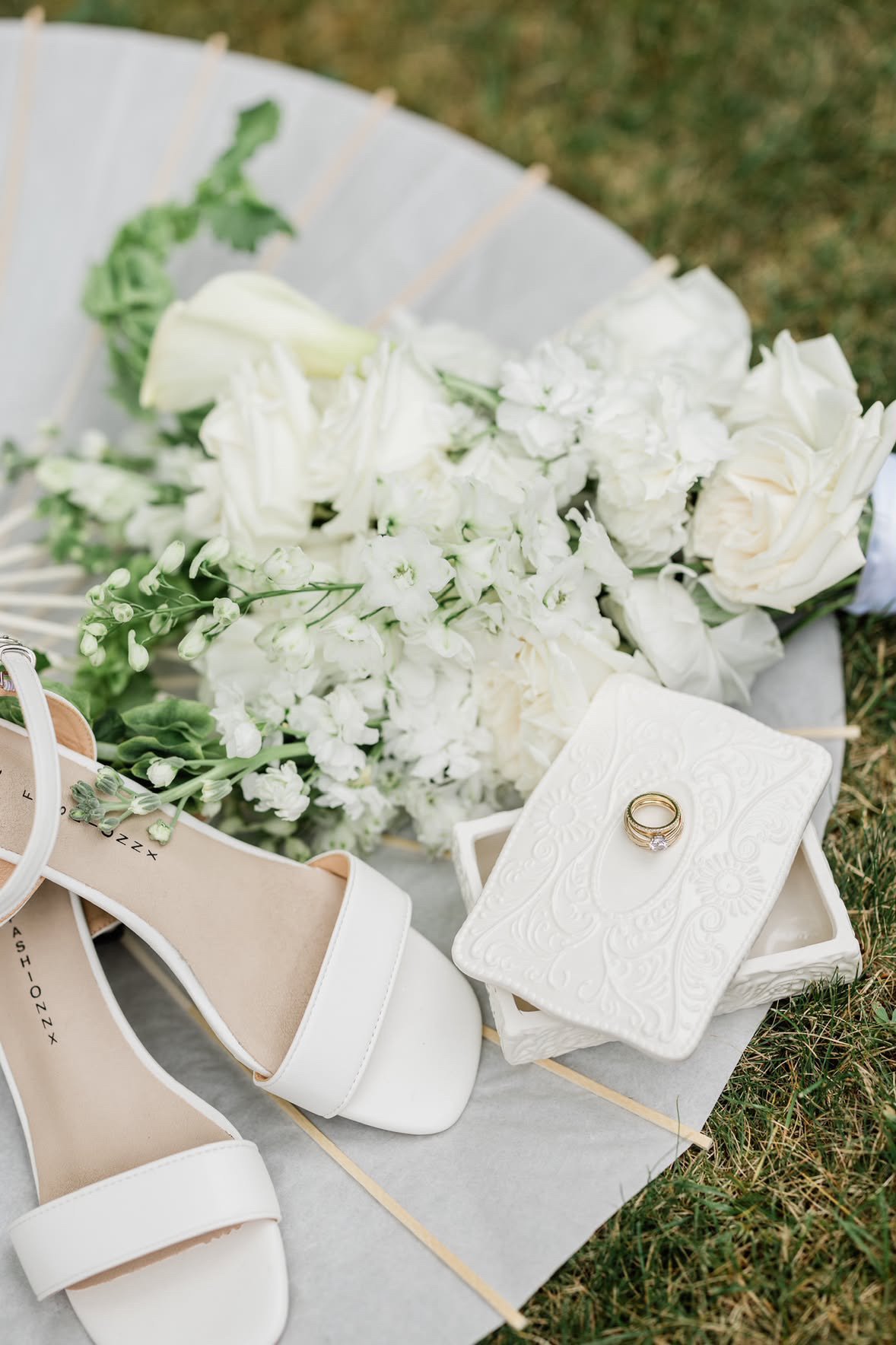A bouquet of white flowers, a wedding ring, a pair of white shoes, and a decorative white tile on grass