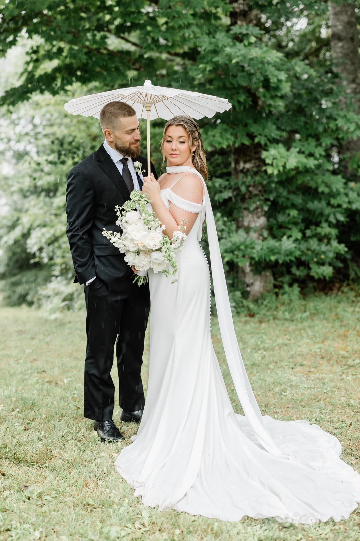 Bride and groom standing outdoors under an umbrella, the bride holding a bouquet of white flowers, wearing a white wedding dress with open shoulders, the groom in a black suit and tie, green foliage in the background, rain falling lightly.