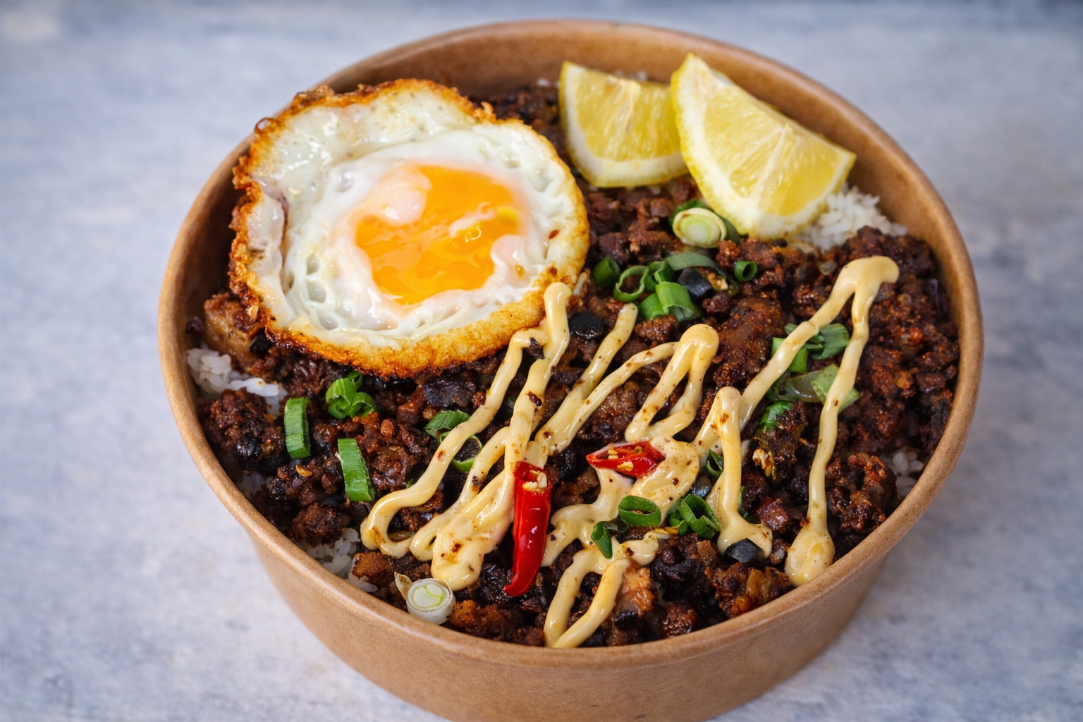 A bowl of rice topped with ground beef, a fried egg, chopped green onions, lemon wedges, and drizzled with a creamy sauce.