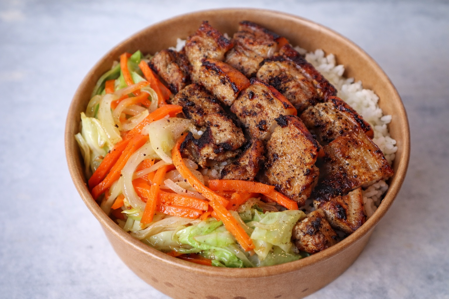 A brown bowl filled with white rice, grilled seasoned chicken pieces, and sautéed mixed vegetables including carrots, cabbage, and celery.
