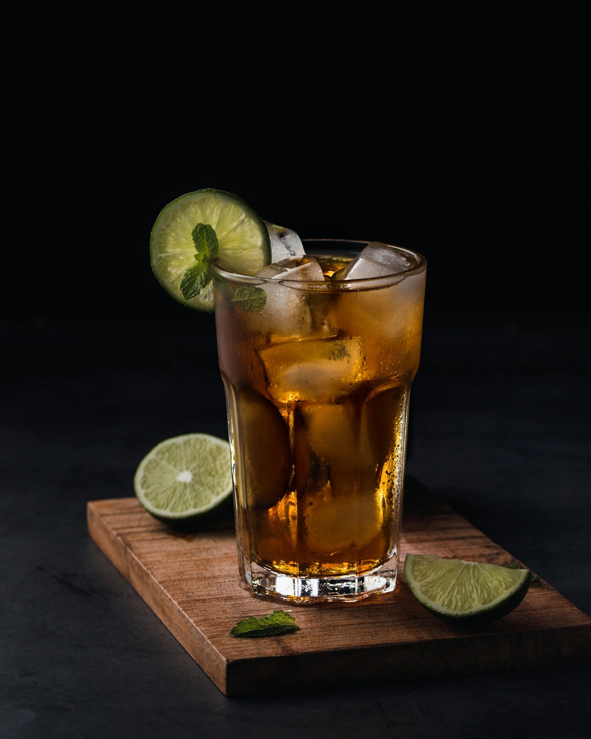 A glass of iced tea garnished with a lime slice and mint, placed on a wooden board with additional lime wedges and mint leaves, dark background.