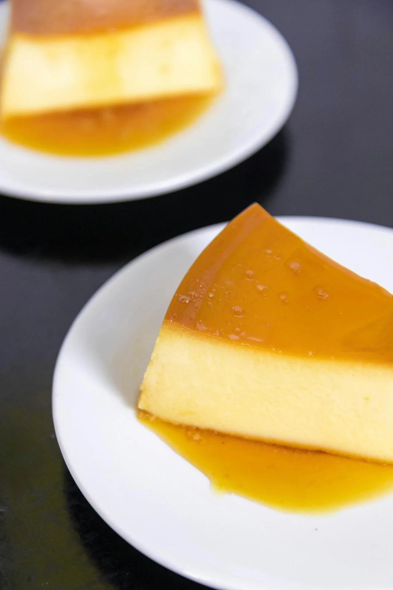 A slice of caramel flan on a white plate with caramel sauce on top, with another piece of flan in the background.