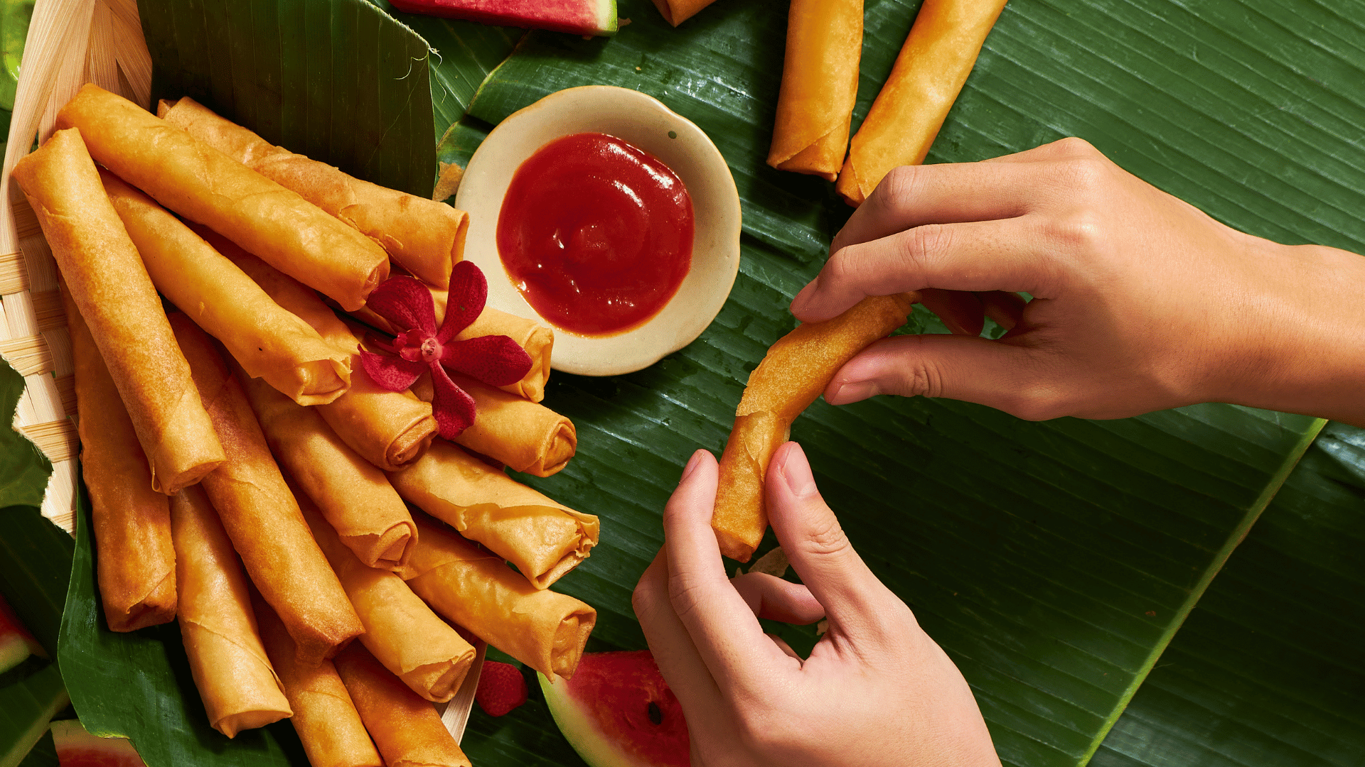 A person rolling a spring roll with fried wrapper, surrounded by more spring rolls and a bowl of red dipping sauce on a green banana leaf table, with slices of watermelon in the background.