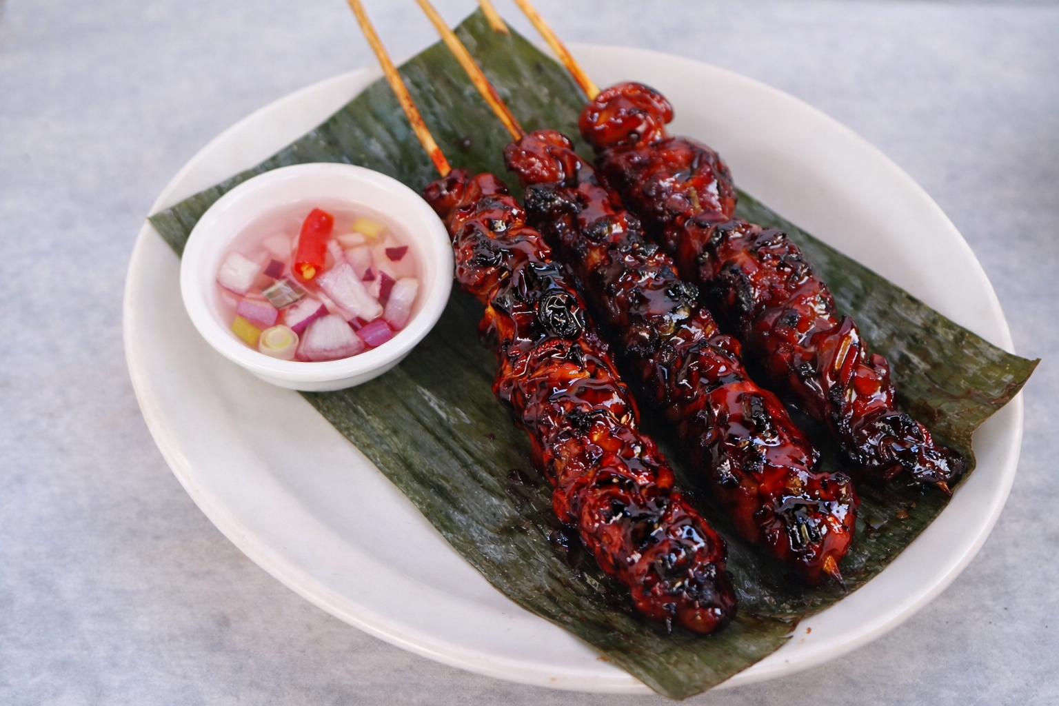 Three skewers of grilled meat with dark glaze served on a banana leaf on a white plate, accompanied by a small bowl of pickled vegetables.