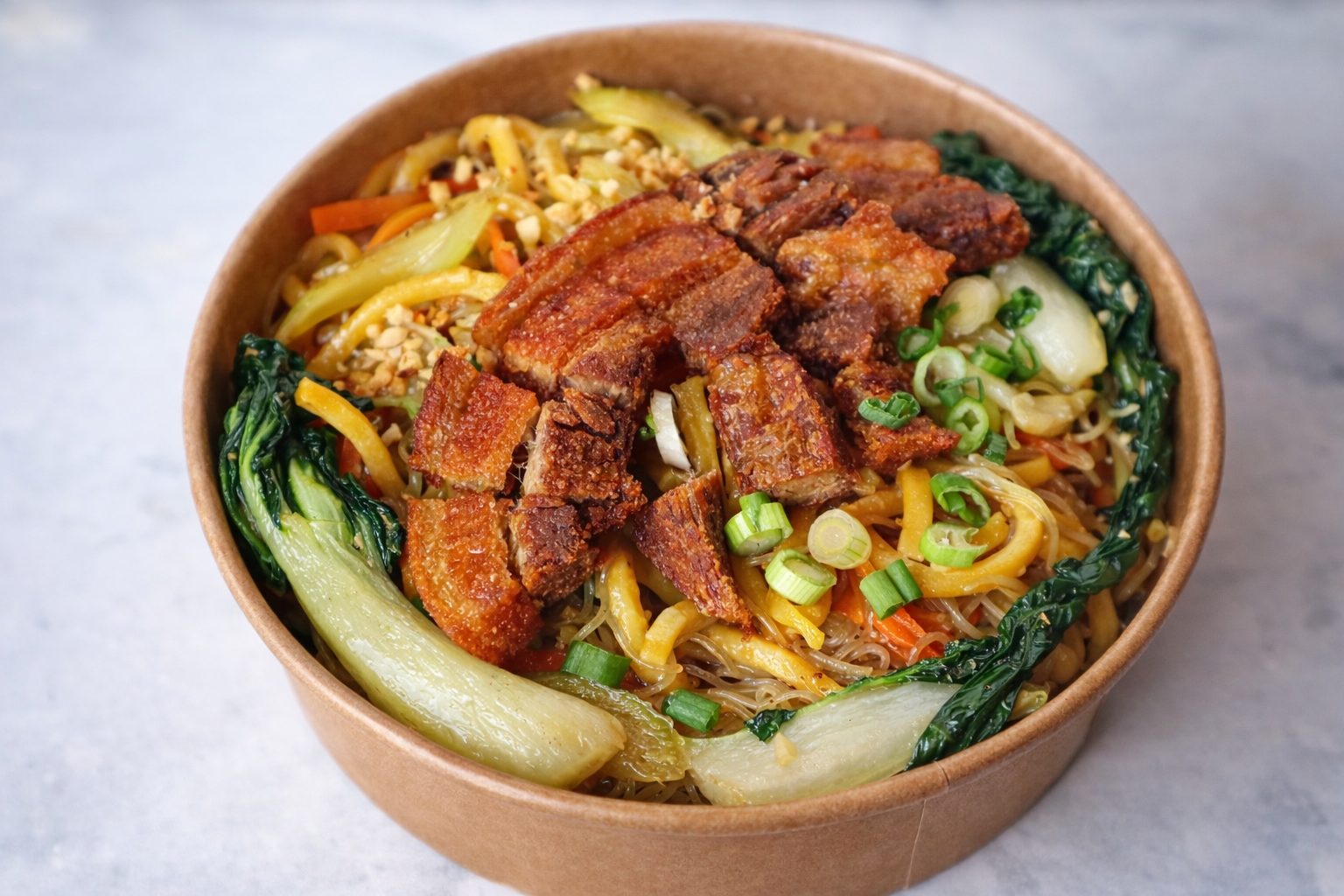Bowl of Asian noodle stir-fry with fried tofu, bok choy, shredded carrots, sliced green onions, and chopped peanuts.