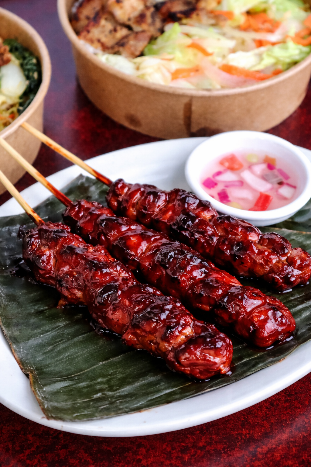 Barbecue skewers with glazed meat served on a banana leaf with a side of pink pickled vegetables in a small white bowl, accompanied by bowls of salad and noodles.