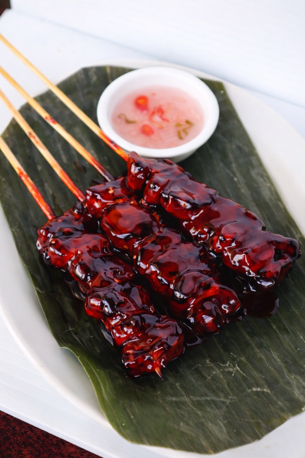 Three skewers of grilled meat coated in dark glossy sauce on a leaf-lined platter with a small bowl of pink dipping sauce