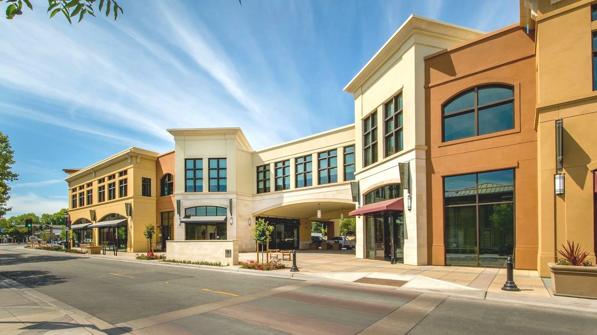 A modern outdoor shopping center with multi-story buildings featuring large windows, storefronts with awnings, and a covered walkway area under the connecting structure. The street in front is empty with crosswalks, and the sky is blue with streaked clouds.
