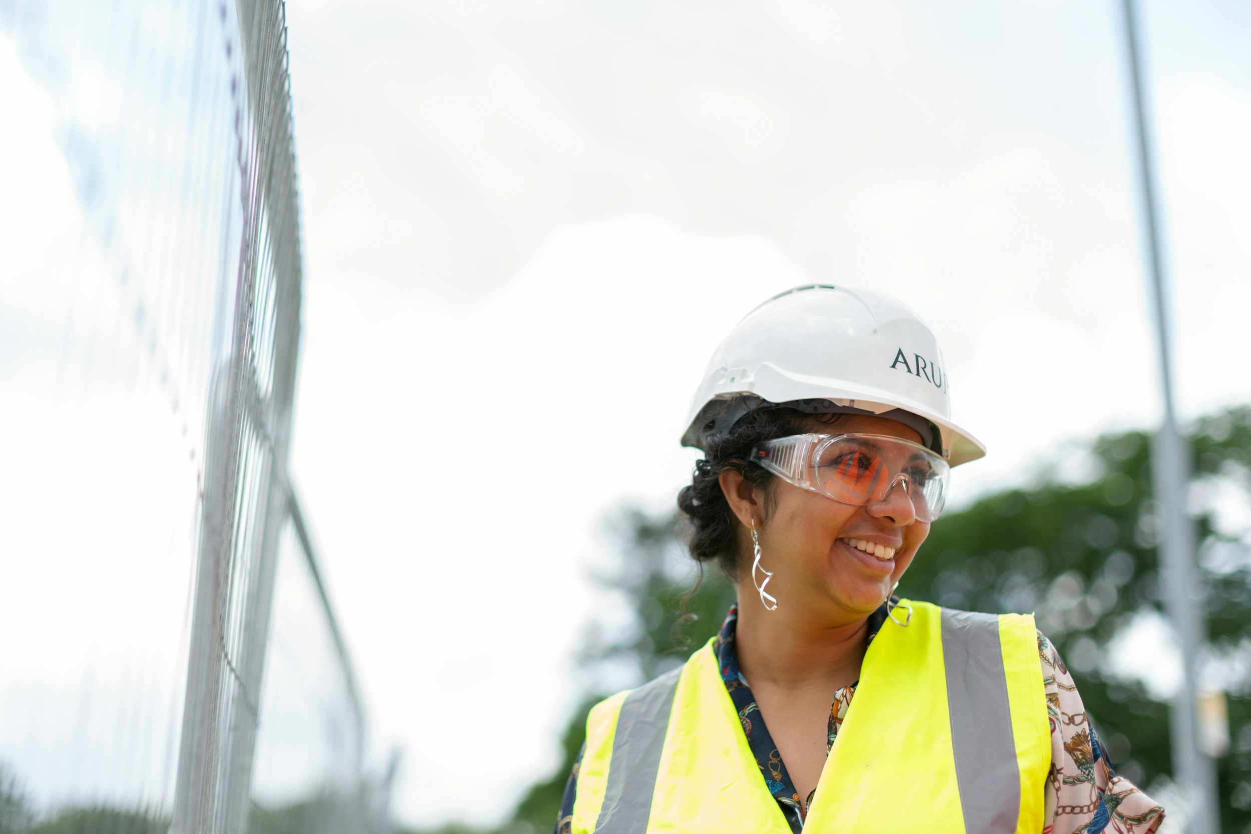 A woman wearing a white safety helmet, safety glasses, and a yellow reflective vest, smiling outdoors near a grid metal structure with a background of trees and cloudy sky.