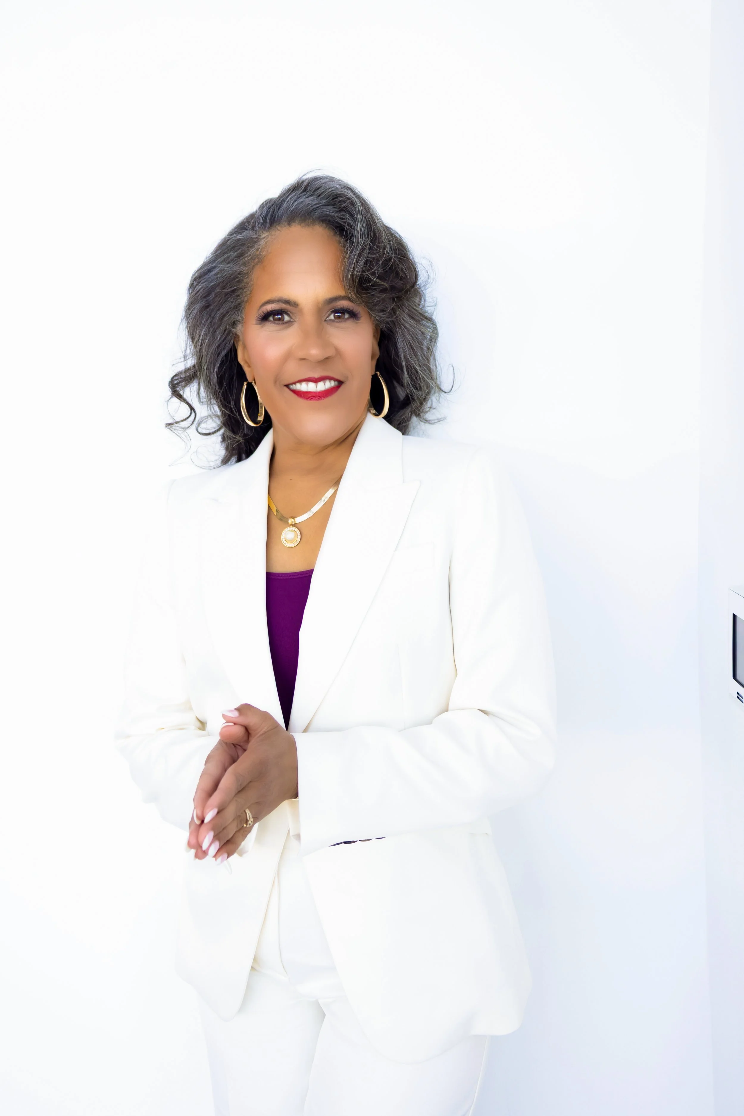 Sharisse Walton wearing a white blazer and pants, gold hoop earrings, and a gold necklace, standing against a white background and smiling.