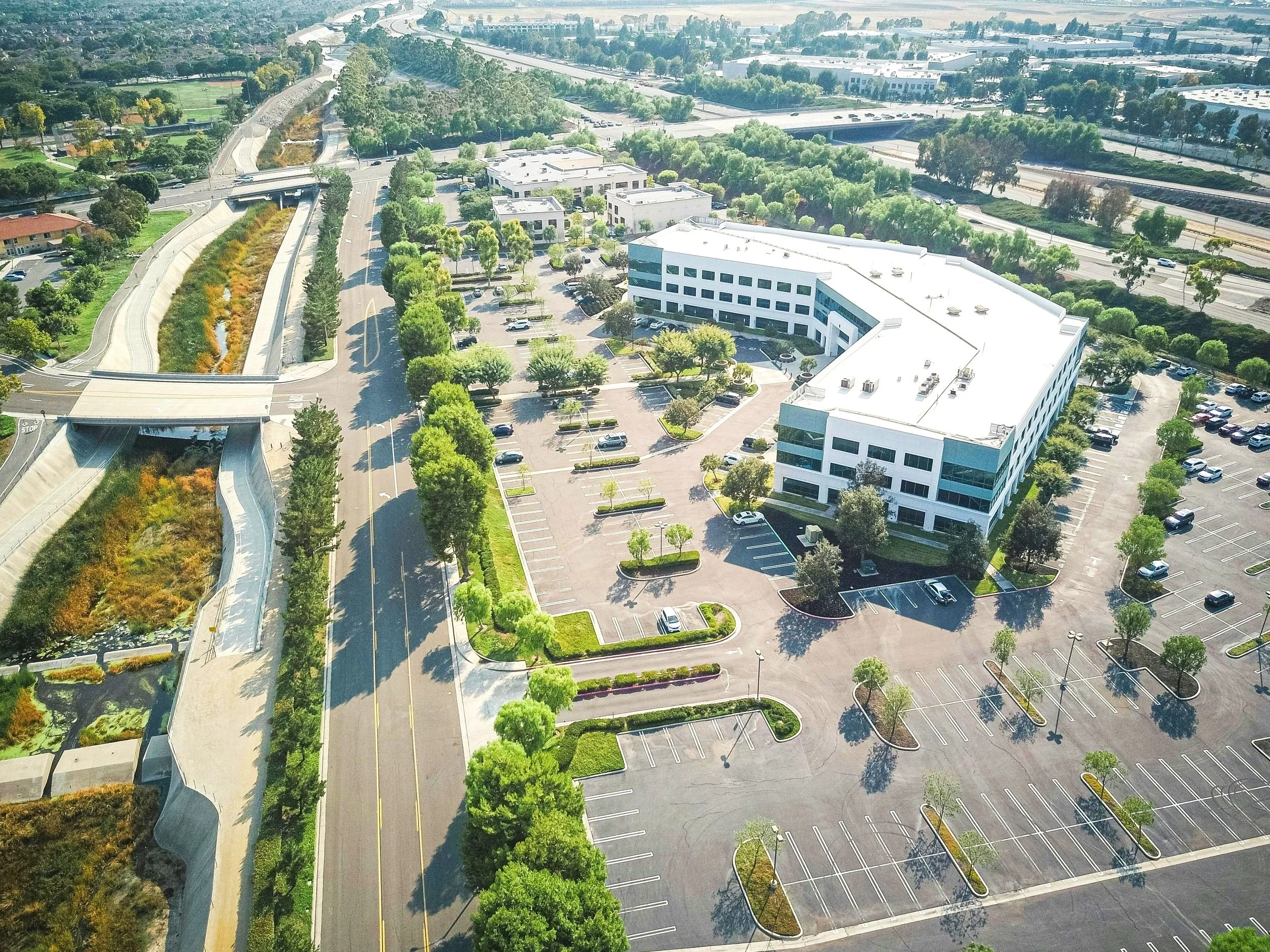 An aerial view of a large office building with a parking lot surrounded by trees, adjacent to highway on a sunny day.