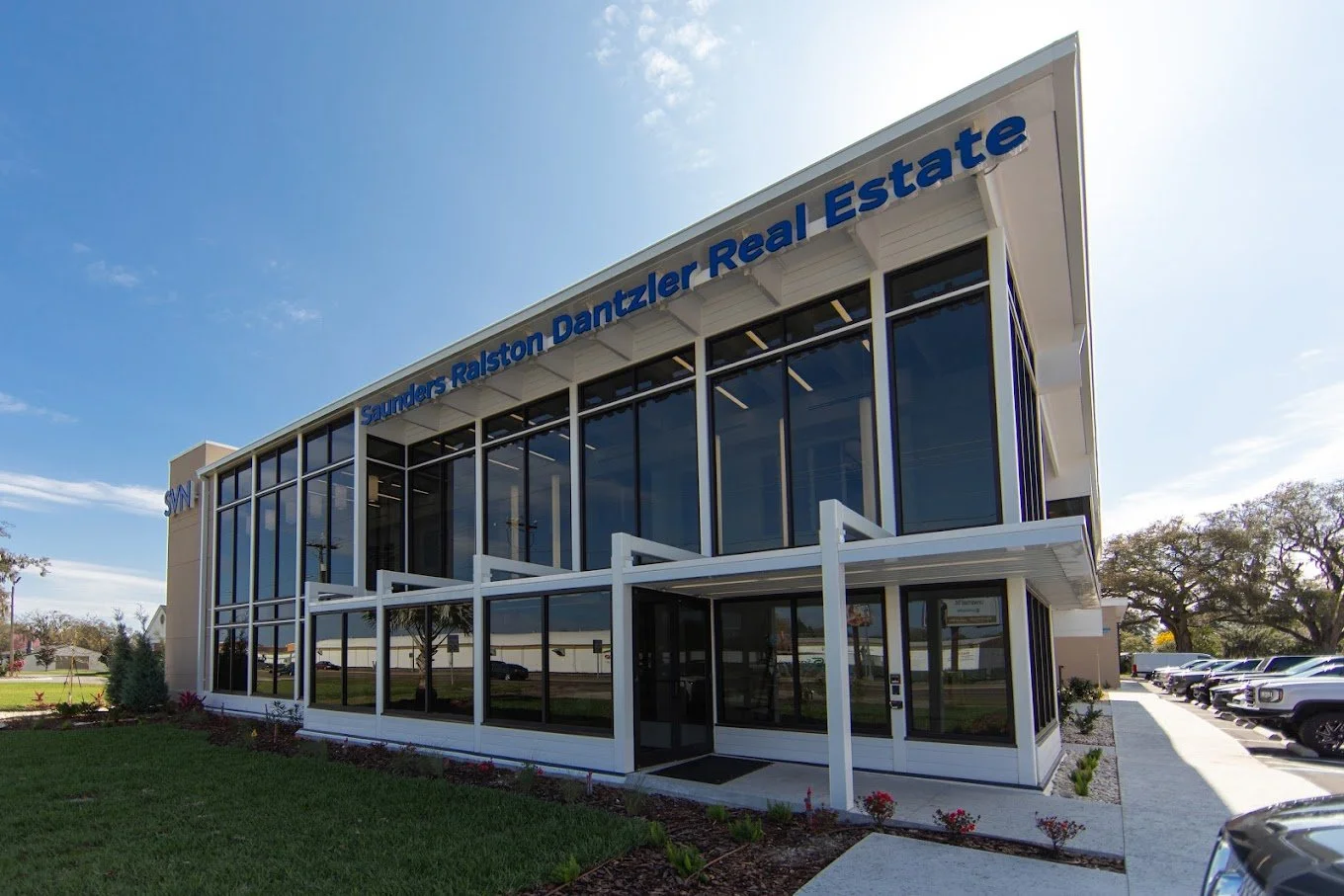 Modern two-story commercial building with large glass windows, a white exterior, and the sign 'Saunders Ralston Dantzler Real Estate' on the top. Some cars parked outside and a lawn with flowerbeds nearby.