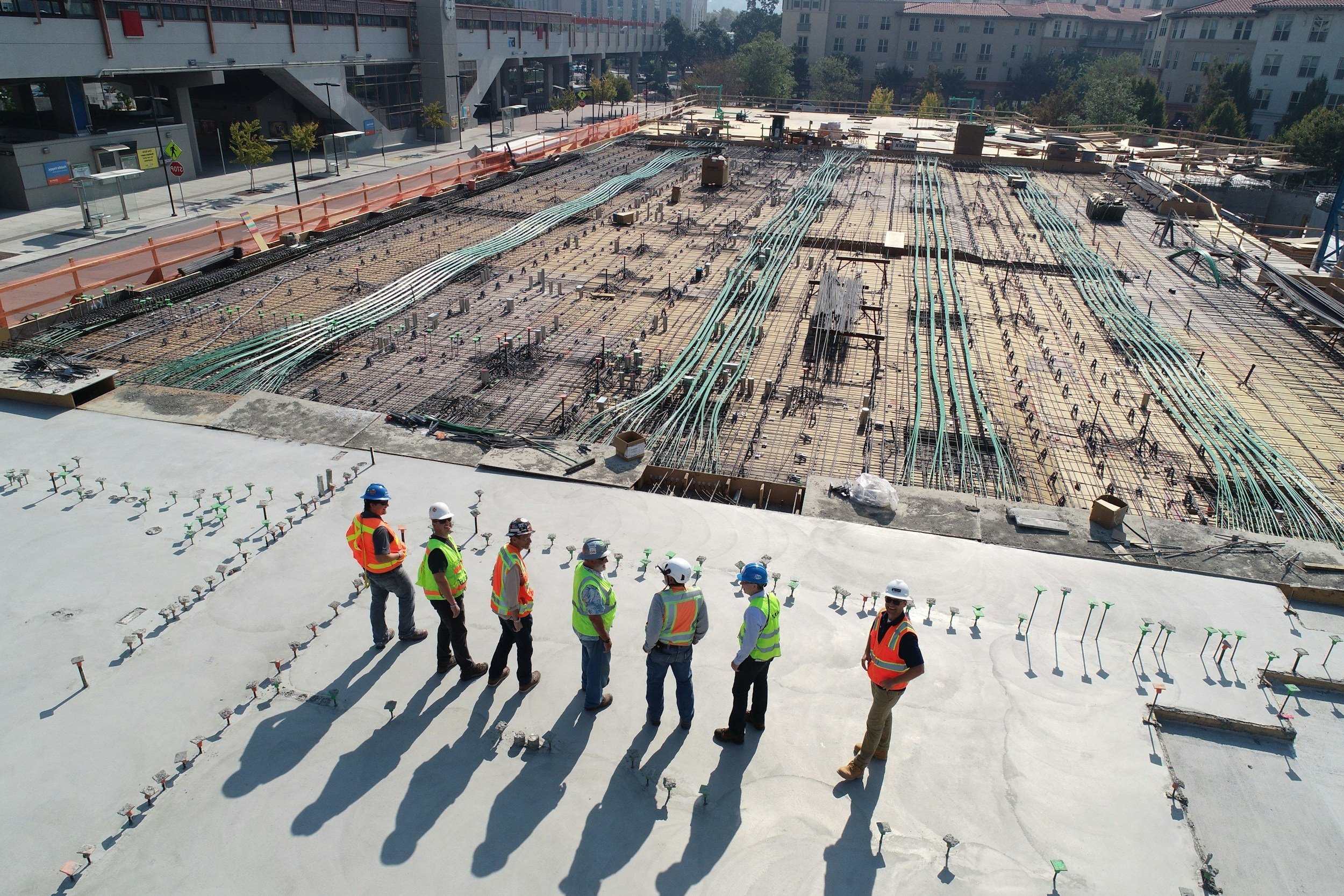Construction workers in safety vests and helmets standing in a line on a building rooftop, overlooking a construction site with exposed rebar, wiring, and construction materials.