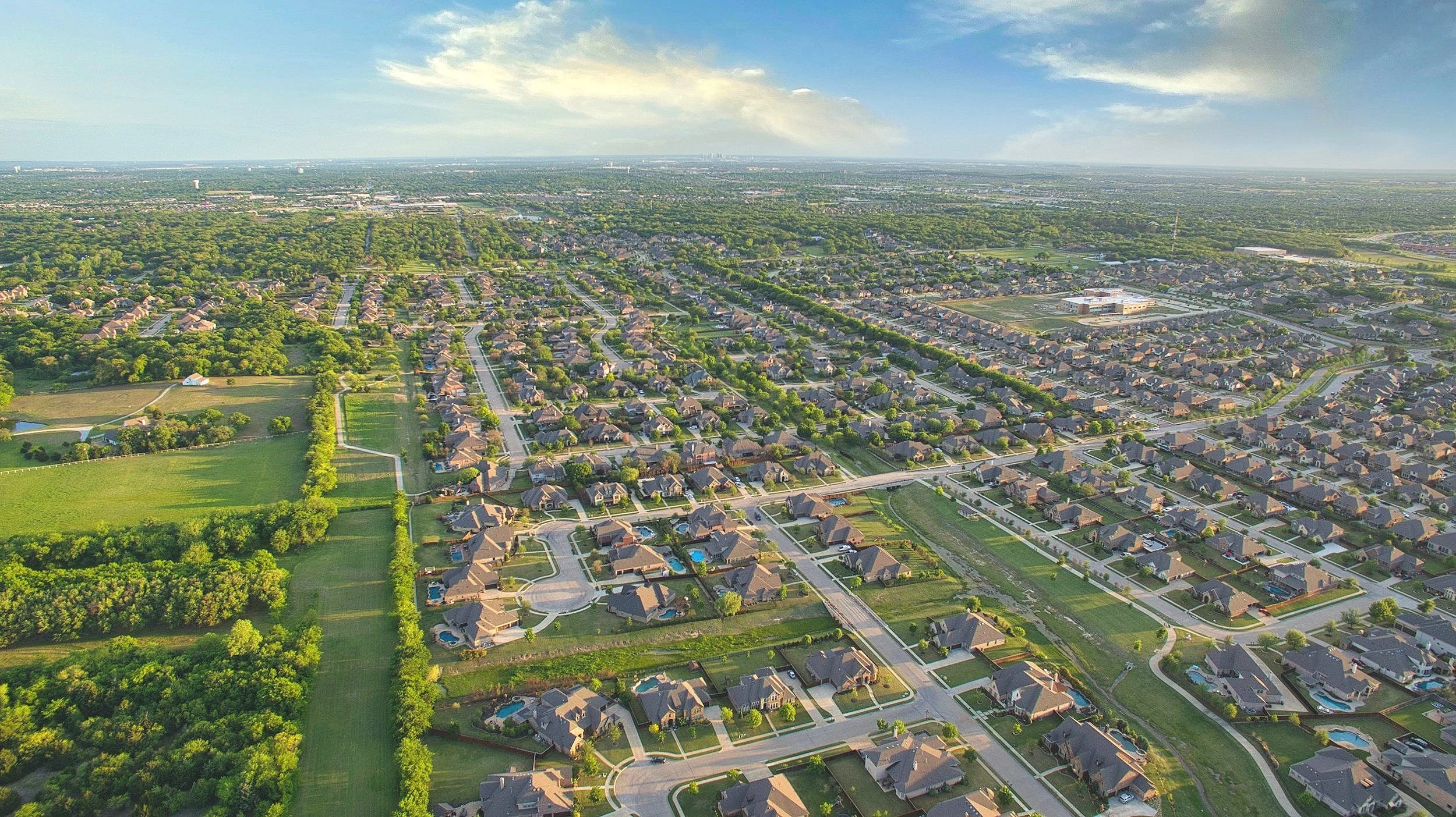 Aerial view of a suburban neighborhood with houses, streets, green lawns, and trees, and a large area of green space and trees on the left side, under a partly cloudy sky during daytime.