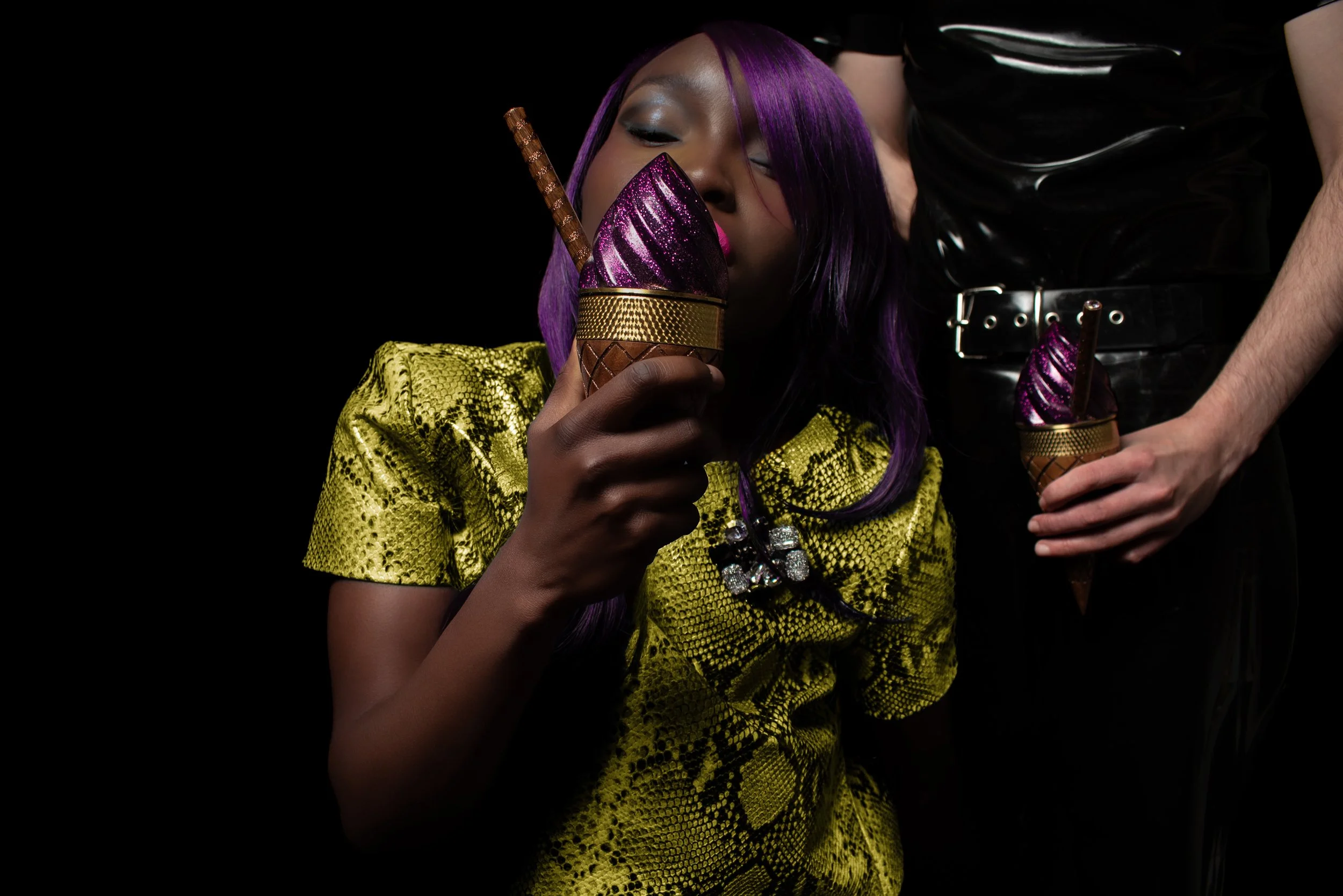 Woman with purple hair drinking from a purple and gold ice cream cone-shaped container holding a chocolate and caramel popsicle, with a person standing beside her holding a similar container with a popsicle.