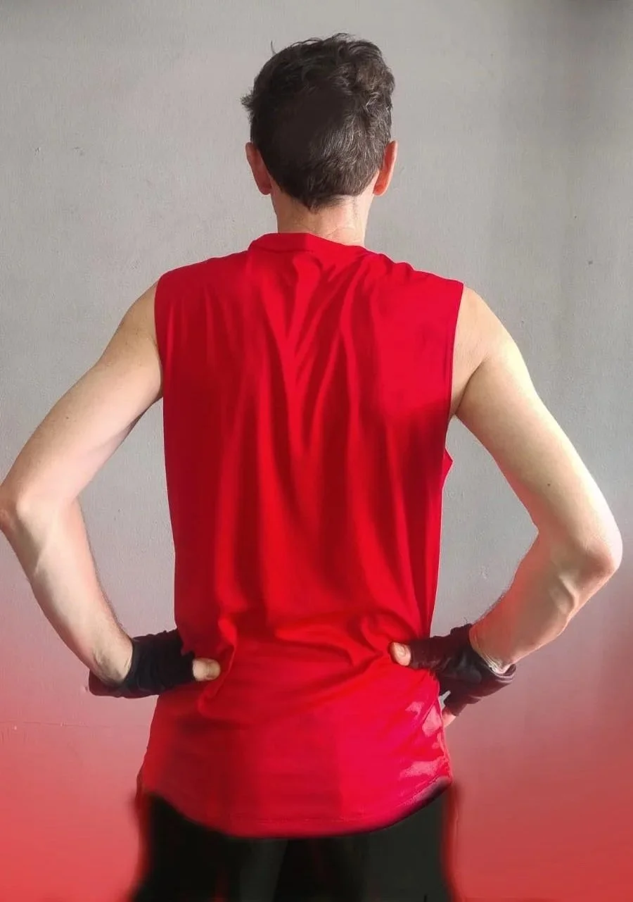 Persona de espaldas con camiseta deportiva roja y guantes negros, de pie con manos en caderas, frente a una pared gris.
