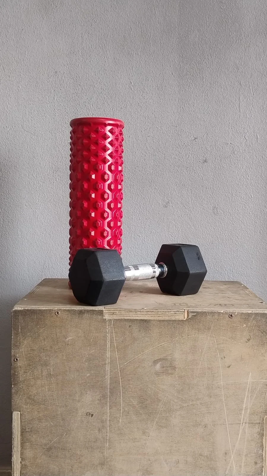Mano sosteniendo una pesa hexagonal negra en una mesa de madera, con un rodillo de masaje rosa y una pared gris de fondo.