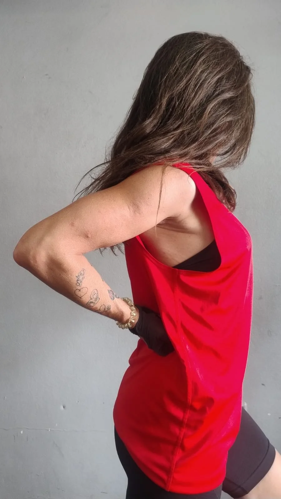 Mujer con cabello castaño, vestido rojo y pantalones cortos negros, vista de perfil con brazo doblado y tatuajes en el brazo.