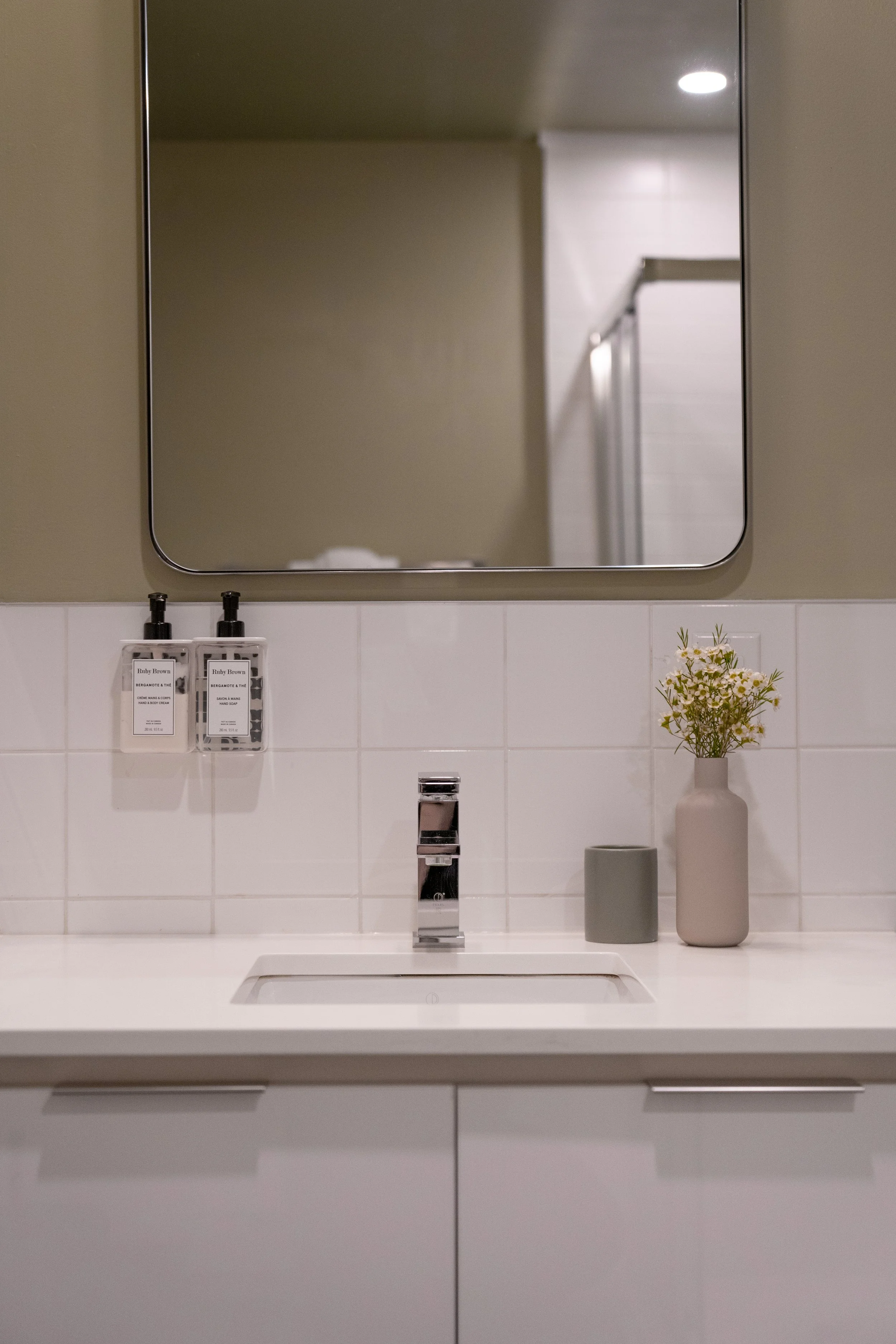 Bathroom sink with a mirror above, soap dispensers on the wall, a gray cup, and a beige vase with flowers on the countertop.