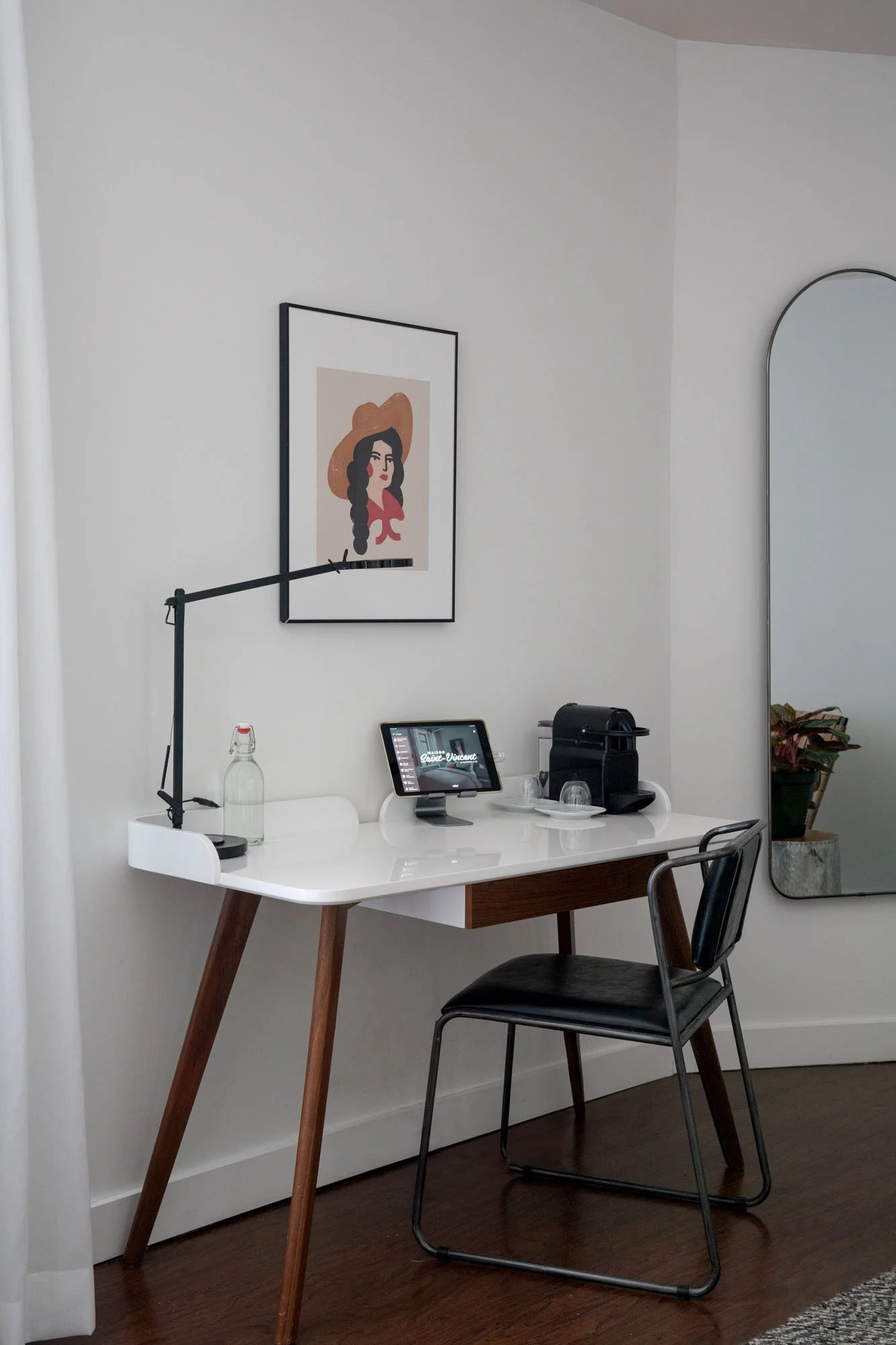 A white desk with wooden legs holding a tablet, a water bottle, a coffee machine, and glasses, positioned in front of a white wall with a framed art print of a woman with dark hair, wearing a large hat and red top, next to a large mirror reflecting a