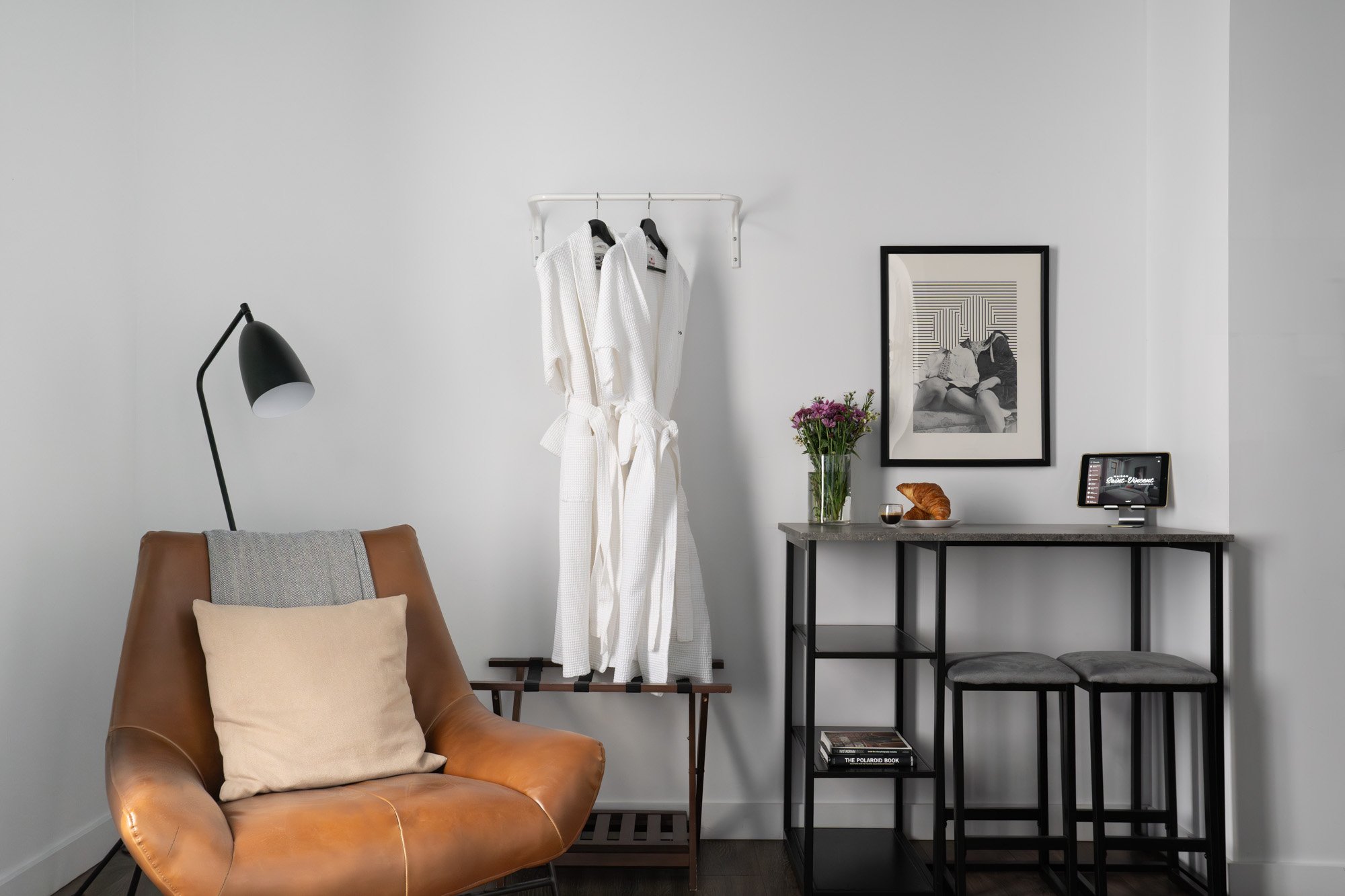 Modern hotel room with brown leather lounge chair, white bathrobes on a rack, black and white framed photograph, vase of purple flowers, croissant, glass of water, small digital device on a table, and two gray stools.