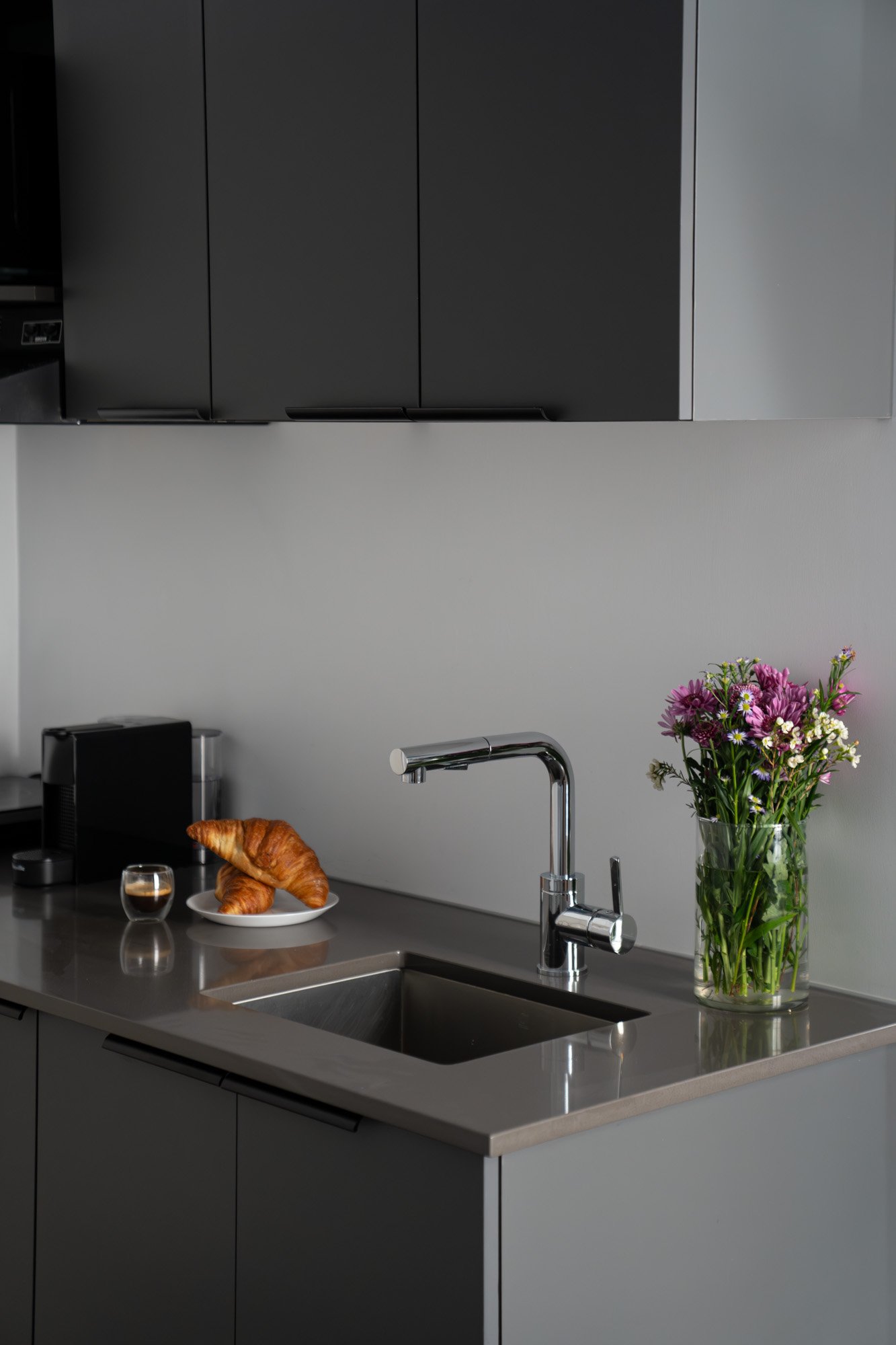 Modern kitchen with stainless steel sink, black cabinets above, a vase of colorful flowers, a plate with croissants, a small glass, and a coffee machine on the countertop.
