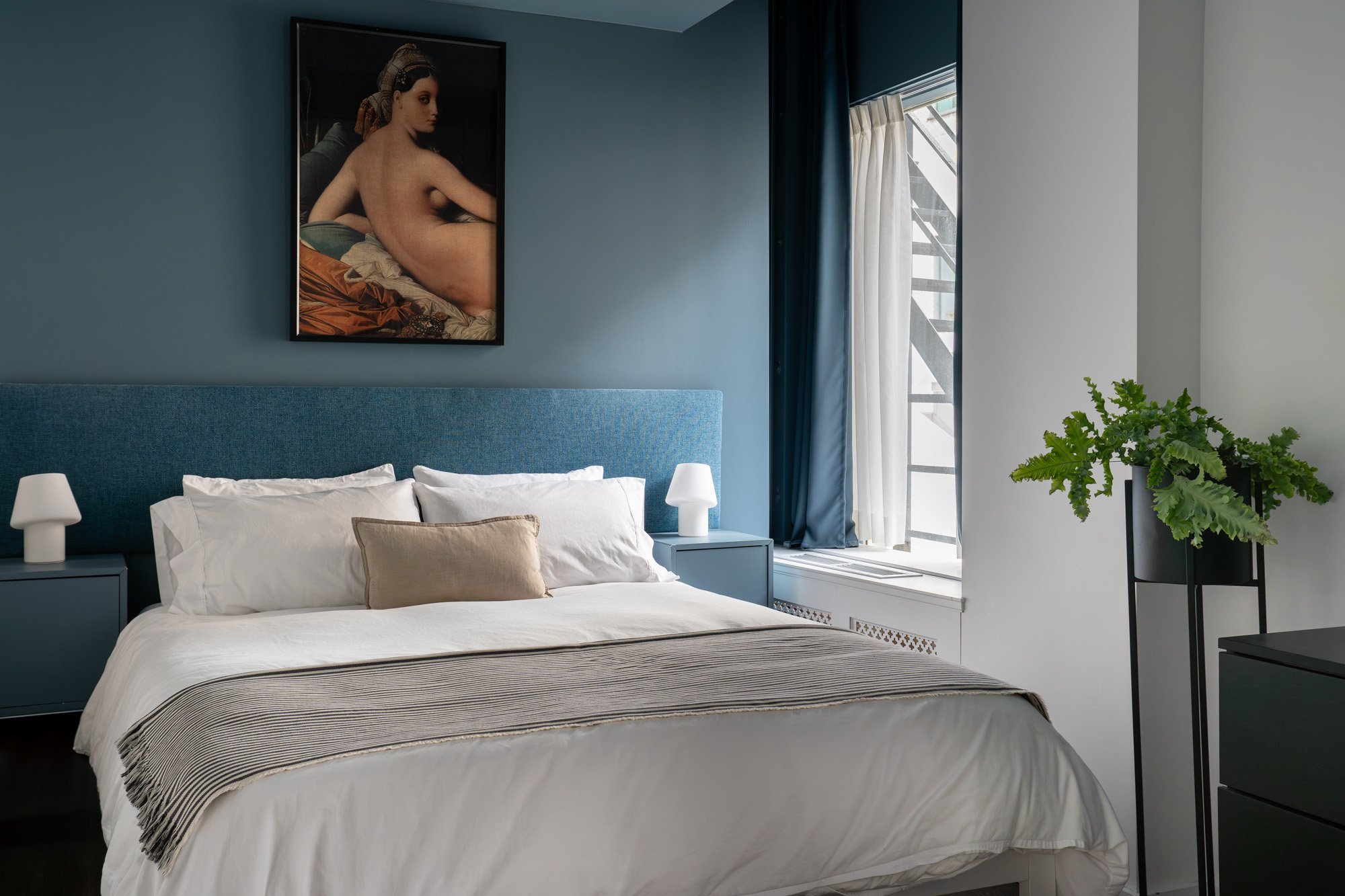 A modern bedroom with a blue upholstered headboard, white bedding, beige pillow, two nightstands with white lamps, a plant on a black stand, and a window with navy blue and off-white curtains. There is a framed painting of a nude woman on the wall ab