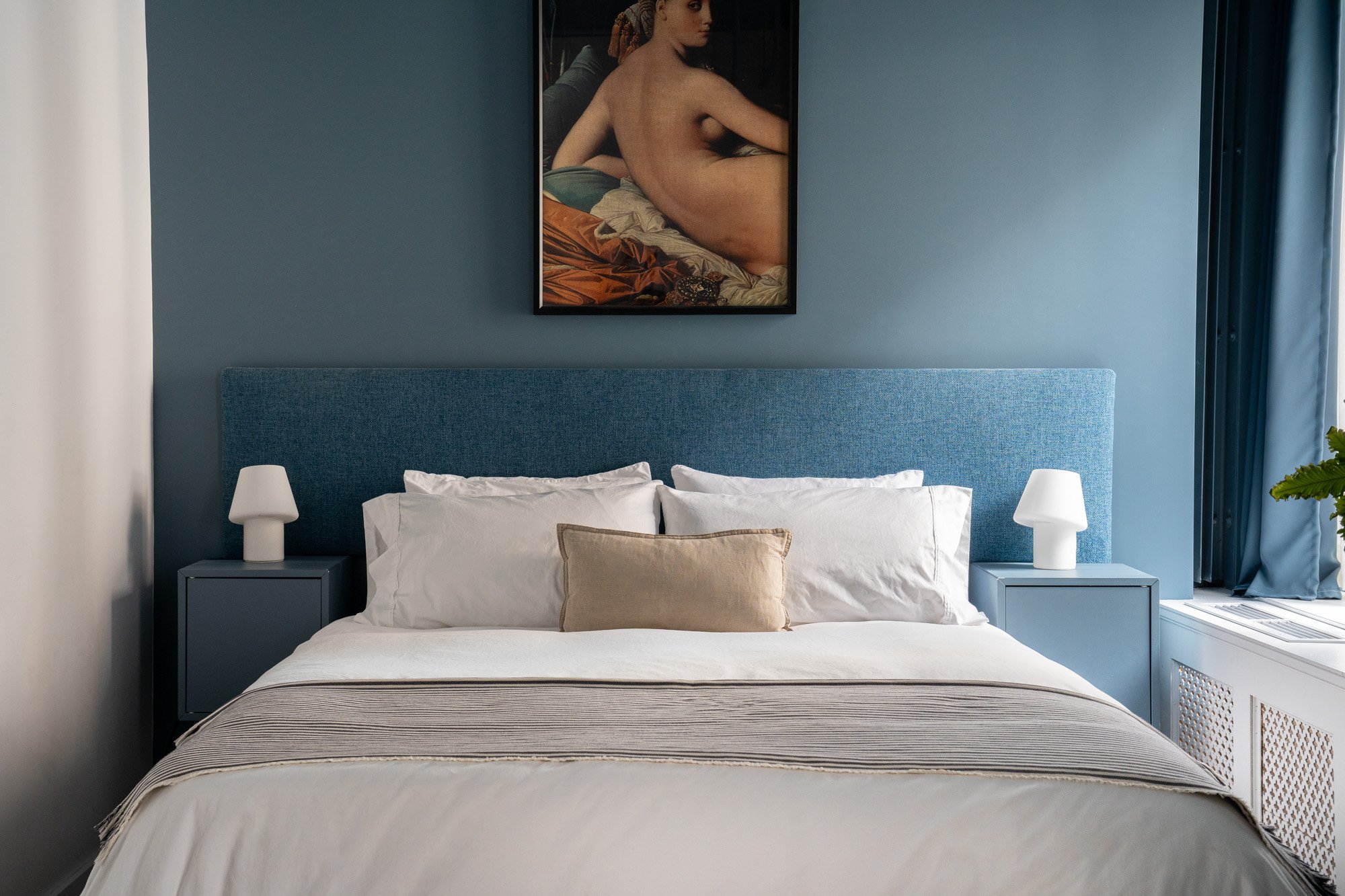 Modern bedroom with blue upholstered headboard, white bedding, beige pillow, blue nightstands with white lamps, blue wall art of a reclining nude woman, and blue curtains.