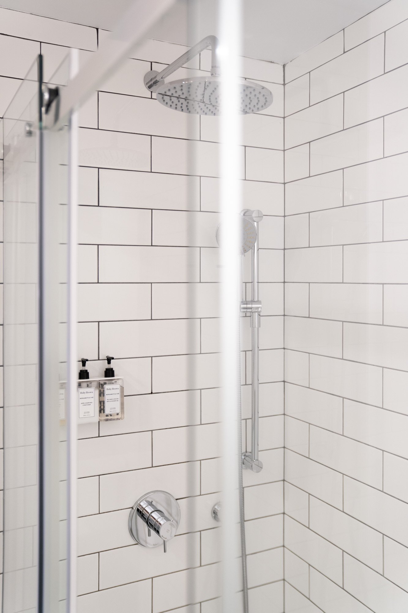 A modern shower with white subway tile walls, a round rain shower head, a handheld shower head, and soap dispensers on the wall.